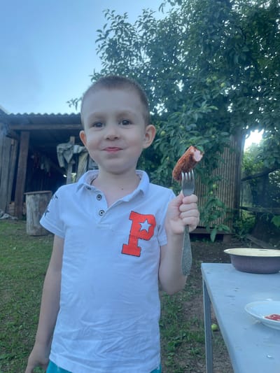 A young boy, appearing to be of Caucasian ethnicity, stands in an outdoor setting. He has light brown hair and is wearing a white polo shirt with an orange letter "P" and a star emblem on the chest. In his right hand, he holds a fork with a piece of cooked sausage on it. He is looking towards the camera with a slight smile.
The background suggests a rural or suburban environment. To the left, there is a rustic wooden structure, possibly a shed or outbuilding, with some items hanging inside and a wooden barrel next to it. Green grass covers the ground. Behind the boy, a tree with green leaves and some small fruits is visible, suggesting it might be an apple tree. The sky is overcast, indicating a cloudy day.
To the right of the boy, a metal table is present. On the table, there is a large bowl and a smaller plate with some red substance, possibly ketchup or sauce, on it. The overall scene depicts a casual outdoor meal or gathering. The location is specified as being in the city of Bormist, Russia.