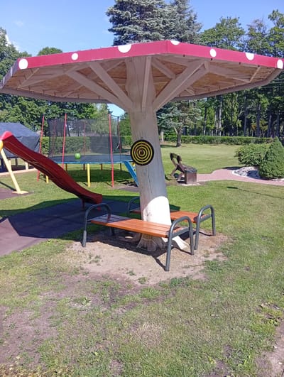 A picturesque scene unfolds in Dirvonėnai, Lithuania, on a bright, sunny day. In the foreground, a charming mushroom-shaped structure with a red and white dotted cap provides shade for a wooden bench. The structure's trunk, made of rough-hewn wood, features a target for darts attached to it. Beside the mushroom, a bright red slide descends onto a rubber mat, part of a children's playground. In the background, a trampoline with a safety net stands ready for use, and a lush green lawn stretches out, dotted with trees and bushes. The overall atmosphere is one of a peaceful, recreational park, ideal for family fun.