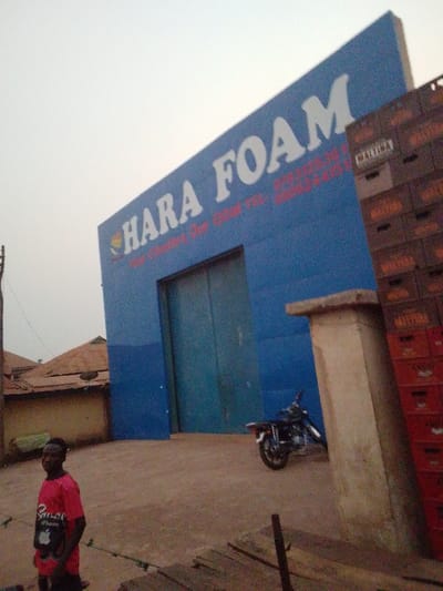 A large blue building with "HARA FOAM" emblazoned on the front stands in the foreground. A large door is visible. A person in a red shirt stands on the left, while a motorcycle is parked near a concrete pillar. Stacked crates labeled "MALTINA" and "AMSTEL" are on the right. A smaller building with a rusted roof is in the background. The sky is overcast.