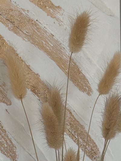 This is a close-up, high-angle, shallow depth-of-field photograph of dried bunny tail grass stalks. The stalks are a light brown or beige color and have a soft, fuzzy texture. They are arranged in a diagonal pattern across the frame, with some appearing closer and more in focus than others. The background is a textured surface that appears to be a wall or canvas. It is primarily white, with textured lines or strokes of a metallic gold or tan color running diagonally, mirroring the direction of the grass stalks. Small flecks of glitter are visible within these gold strokes. The lighting is soft and diffused, suggesting an indoor setting with no strong shadows. There is no discernible time of day or weather, and the location context of Cluses, France, provides no specific visual cues within the image itself. The overall mood is calm and decorative, with an emphasis on natural textures and subtle metallic accents.