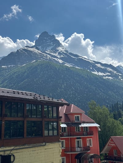 The image shows a landscape scene with a mountain and buildings in Dombay, Russia. The mountain is covered in snow and trees, with a bright blue sky and fluffy white clouds above. There are several buildings in the foreground, including a red building with balconies and windows, and a wooden building with large windows. A sign on the red building reads "Отель Кампус" (Hotel Kampus). The time of day appears to be daytime, with clear weather. The scene evokes a sense of natural beauty and a mountain resort atmosphere.