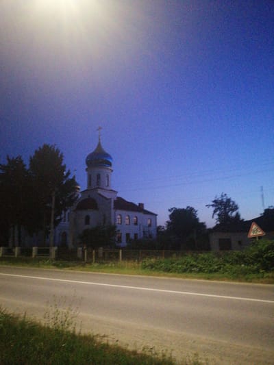 The image captures a view of a road alongside a cluster of buildings and trees under a twilight sky. In the background, a white church with a blue dome and a cross is prominently visible, its architectural style suggesting it could be an Eastern Orthodox church. Adjacent to the church are other buildings and dense foliage. To the right, a triangular road sign with an emblem is partially obscured by greenery. The foreground features a paved road with a white lane marker and a strip of grass and weeds along the side. The lighting suggests it is either early morning or late evening, with a bright light source, possibly the sun or artificial lighting, dominating the upper left portion of the sky. The scene is devoid of any people or animals, focusing solely on the architectural and natural elements of the location. The location is identified as being in the city of Lanivtsi, Ukraine.