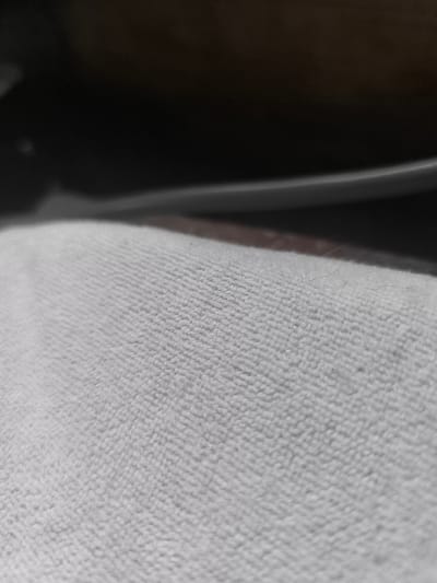 The image is an extreme close-up of a textured, light-colored fabric, possibly a towel or blanket. The fabric has a dense, looped pile that creates a ridged pattern. The lighting suggests the fabric is illuminated from the side or top, casting subtle shadows that emphasize the texture. In the upper portion of the image, a darker, out-of-focus area is visible, which could be furniture or another surface. There is no discernible information about the city of Balezino, Russia, or any people, actions, or interactions in the image.