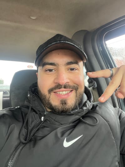 A male individual is captured in a selfie taken within a vehicle in Oued Jdida, Morocco. The subject, a man with dark hair, a full beard, and a mustache, is wearing a dark-colored baseball cap and a black puffer jacket featuring a white Nike swoosh logo on the left chest. He is smiling directly at the camera. His right hand is raised, with the index and middle fingers extended downwards towards his left shoulder/chest; a dark-colored ring is visible on his right ring finger. The vehicle's interior shows a light-colored headliner and a dark fabric car seat behind the man. A seatbelt mechanism is visible on the right side of the frame. Through the vehicle's windows, raindrops are evident, and the external environment appears overcast and indistinct. A small red and white circular sticker is partially visible on the upper left of the window area behind the subject.