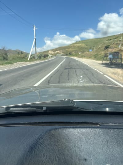 The image shows a view from inside a car looking out through the windshield at a winding road. The road is paved and has white lane markings. There are power lines and a utility pole on the left side of the road. A green, grassy hillside is visible on the right side, with a few scattered bushes and trees. In the distance, there appears to be a small, weathered structure or cart on the side of the road. The sky is blue with scattered white clouds. The car's dashboard and windshield wipers are visible in the foreground. The scene suggests a rural or semi-rural location, possibly along a highway or main road in a mountainous or hilly region. The location is identified as Zangakatun, Armenia.