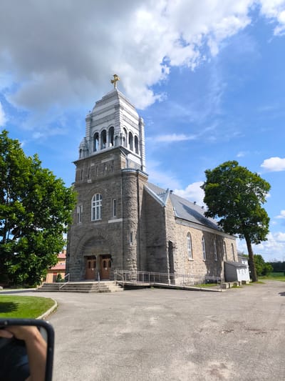 The image shows a stone church with a tall bell tower topped with a golden cross. The church has arched doorways and windows. A ramp provides access to the entrance. Green trees flank the church, and the sky is blue with scattered clouds. The scene is set in Saint-Philippe-d'Argenteuil, Canada, as indicated by the contextual information. A hand holding a phone is partially visible in the lower left corner, suggesting the image was taken by someone using a mobile device.