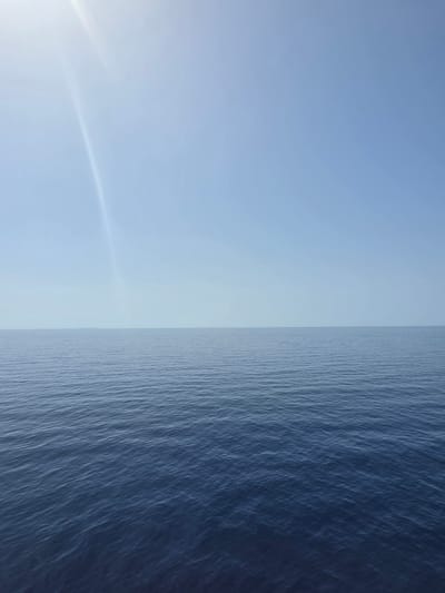 The image shows a wide expanse of calm, dark blue ocean under a clear, pale blue sky. The horizon line is sharp, dividing the water from the sky. A faint, bright streak of light, possibly a sunbeam or lens flare, is visible in the upper left quadrant of the sky. The water's surface is gently rippled, indicating minimal wave activity. There are no people, boats, or landmasses visible in the frame, suggesting an open sea environment. The scene is devoid of any urban elements or specific landmarks that would place it within the city of Santa Ponça, Spain.