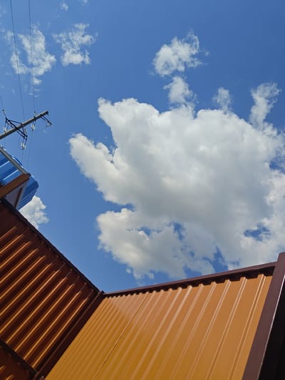 The image, captured in the city of Shkurupiivka, Ukraine, depicts a bright, clear sky with scattered white cumulus clouds. In the foreground, two sections of a corrugated metal roof are visible, one a rich copper color and the other a burnt orange hue, meeting at an angle. To the left, a utility pole with power lines and insulators stands against the sky, partially obscuring a blue-colored structure, possibly a portable toilet or storage unit. The perspective is from below, looking upwards.