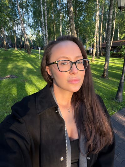 A woman with long brown hair and glasses is posing for a selfie outdoors. She is wearing a black jacket over a grey shirt. The background shows a green lawn and birch trees in a park. The location is likely Polivanovo, Russia.