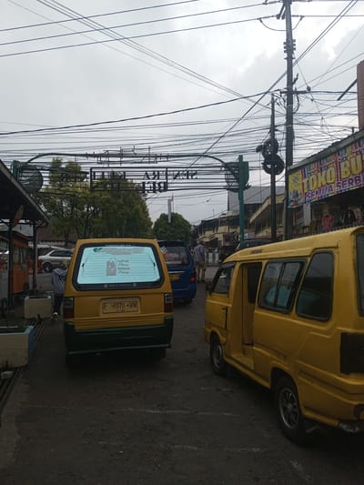 The image displays an urban street scene under an overcast sky. The visual evidence, including Indonesian language text on vehicle stickers and commercial banners, and an Indonesian-format license plate, contradicts the provided location of Houaythat, Laos, strongly indicating a location within Indonesia.
In the foreground, two yellow minivans are prominent. The leftmost minivan, with a green lower bumper, displays an Indonesian license plate "F 1976 WW" with an "11-26" expiry. Its rear window features a sticker reading "HARUS PULANG SEBELUM HILANG" and "Panca Tunggal." A person wearing a grey hoodie, dark pants, and light shoes is partially visible walking from left to right behind this vehicle. To the right, a second yellow minivan, showing signs of wear and rust, has its passenger-side door open.
In the mid-ground, a blue minivan with a partially visible license plate "D 8092" is positioned behind the first yellow minivan. Further left, a silver sedan is visible behind roadside structures. The road surface is dark, worn asphalt.
The right side of the street features a building with a corrugated roof, adorned with a large banner displaying Indonesian text ("TOKO BUS..." and "SEMUA SERBA...") and Arabic script, beneath which clothing is visible for sale. Numerous black electrical and communication wires crisscross overhead, supported by multiple utility poles, one of which is prominent on the right with insulators and coiled wires. An overhead metal archway with a reversed sign (likely Indonesian) spans the street. A green traffic light is visible on the right. Green trees are present in the background, and additional indistinct human figures are visible further down the street.