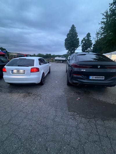 A black BMW X6 and a white Audi A3 are parked side-by-side in a parking lot. The BMW's license plate reads "OB MMMMMM" and the Audi's reads "EH MMMMMM". The photo was taken in Zlatna Panega, Bulgaria.