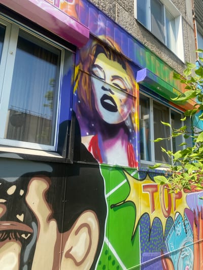 This is a close-up, medium-angle, outdoor shot of a building facade adorned with vibrant graffiti. The graffiti depicts a stylized portrait of Marilyn Monroe, with her eyes closed, her mouth slightly open, and her hair a blend of yellow, orange, and purple. She is wearing a red dress. To the left and bottom of the image, another section of graffiti shows a partial view of a person's face, featuring an eye and part of the nose and mouth, rendered in black, brown, and beige tones. The building itself has white-framed windows and a grey, textured wall. Sections of the building's exterior are painted with brightly colored panels featuring comic-book-style lettering and abstract designs, including the word "TOP" in red lettering on a yellow background. A green leafy branch extends into the right side of the frame, partially obscuring the graffiti. This image was taken in the city of Achinsk, Russia.