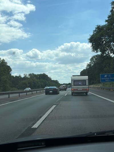 The image captures a view from inside a vehicle traveling on a highway, likely the A3 in the vicinity of Hünxe, Germany, as indicated by the highway sign for Dinslaken-Nord. The sky is a bright blue with scattered white clouds. On the right side of the highway, a blue sign with white lettering reads "A 8 Dinslaken-Nord 1000 m." A line of trees and foliage forms a dense border along the right side of the highway. On the left side of the highway, a metal guardrail separates the lanes from the adjacent terrain.

The highway is moderately busy with several vehicles. In the foreground, the dashboard and windshield wiper of the capturing vehicle are visible. A black sports car is in the right lane, and a white caravan is in the left lane ahead of it. Further ahead, other cars are visible in both lanes, indicating traffic flow. The road surface appears to be asphalt, with clear lane markings. The overall scene depicts a typical highway journey under a partly cloudy sky.