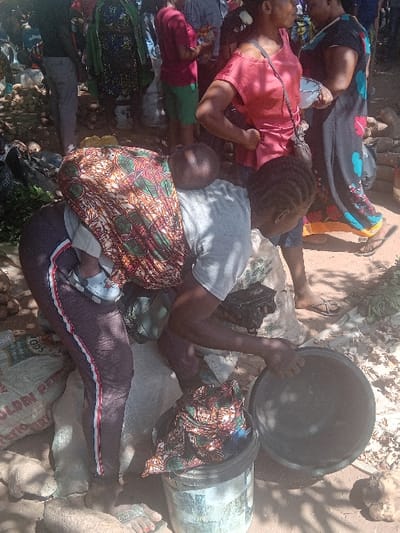 The scene depicts an outdoor market area in Gurku Gwari, Nigeria, under daylight conditions. In the foreground, a woman is bent forward, with an infant secured to her back by a colorful, patterned fabric wrap. The infant wears light blue sandals. The woman is dressed in a grey t-shirt and dark leggings with red and white stripes along the outer seams. Her right hand holds a dark, empty circular bowl over a light-colored plastic bucket that contains a piece of patterned fabric. To her left, a large, partially visible white sack marked "GOLDEN R..." is on the ground. Multiple root vegetables are scattered on the uneven, light brown dirt ground around the woman's feet.
In the midground and background, several other individuals are present, indicating a crowded environment. One woman stands to the right, facing away from the camera, wearing a red off-shoulder top and dark pants; she has a dark shoulder bag. Another woman in a dark, patterned long dress is also visible. Further back, a person in a red t-shirt and green shorts is observed holding items. The ground is consistently dirt with scattered debris and organic matter.
