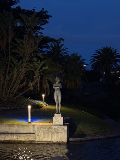 At night, in Milnerton, South Africa, a statue of a nude person stands on a concrete base near a body of water. The scene is illuminated by ground-level lights, with trees and foliage surrounding the statue. The sky is a dark blue.