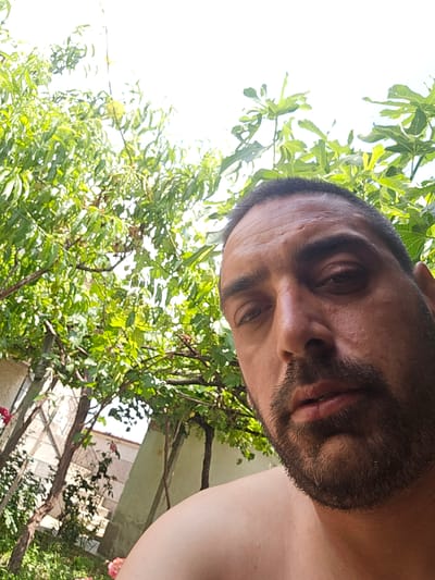 This is a selfie taken outdoors in the city of Brezovo, Bulgaria. The image features a man's face in the foreground, with a beard and short dark hair. He is looking directly at the camera. Behind him, lush green foliage from trees and plants is visible, suggesting a garden or natural setting. The bright sky is visible through the leaves, indicating it is daytime. Part of a building with a textured wall and some pink flowers are also present in the background to the left. The perspective suggests the photo was taken from a low angle, looking up at the man and the surrounding greenery.