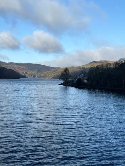 This image depicts Ladybower Reservoir, situated within the High Peak district of the United Kingdom, under clear daytime conditions. The foreground and midground are dominated by the dark blue, rippled surface of the reservoir water. Wooded hills, predominantly showing bare deciduous trees, rise from the water's edge on both the left and right sides, extending into the background. On the right side of the reservoir, a distinct promontory extends into the water, hosting a stone-built house with a dark grey or black roof, partially obscured by trees. The sky is bright blue with scattered white cumulus clouds, and sunlight is visible illuminating portions of the distant hills and clouds. No individuals or watercraft are present or discernible in the frame.