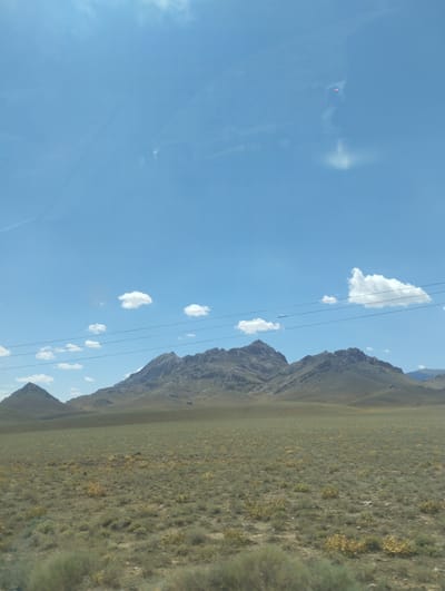 This is a photograph of a vast, arid landscape under a bright blue sky dotted with scattered white clouds. In the foreground, a dry, grassy plain stretches out, sparsely covered with low-lying, scrubby vegetation in shades of green and yellow. The terrain appears relatively flat, leading towards a range of imposing, rugged mountains in the middle ground. These mountains are rocky and arid, with visible textures and formations suggesting erosion. A set of power lines with several wires runs horizontally across the mid-section of the image, stretching between unseen poles. The sky is clear and dominates the upper half of the frame, with a subtle reflection or glare visible in the upper left portion, possibly from a window or lens. The overall impression is one of a remote, natural environment, typical of a desert or steppe region. The location is described as Asmansay, Uzbekistan.