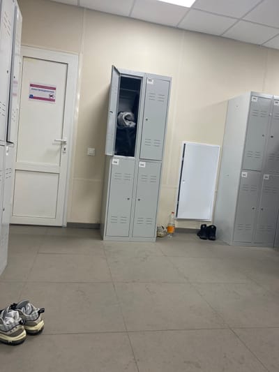 The image displays an interior view of a changing area located in Nikol'skoe, Russia. The room features light beige walls, a light grey tiled floor, and a white suspended ceiling with visible grid lines and integrated lighting.
On the left, a white interior door with a silver lever handle is visible. A rectangular sign with Russian text "ЗОНА ПЕРЕОДЕВАНИЯ" (Changing Area) and two graphic symbols is affixed to the door. Directly adjacent to the door, a stack of grey metal lockers is present, comprising four individual units arranged in two columns and two rows. The upper-left locker, numbered 555, is open, revealing dark fabric or clothing inside. The remaining three lockers in this stack are closed and numbered 556 (bottom left), 553 (top right), and 554 (bottom right). Each locker door features horizontal ventilation slats.
To the right of the central lockers, a white rectangular access panel or small door is partially ajar on the wall. Further to the right, another stack of grey metal lockers is partially visible, with doors numbered 551, 552, 550, and 549, all appearing closed.
On the floor, in the foreground to the left, a pair of light-colored athletic shoes with thick soles are positioned. Near the central locker unit, an orange plastic bottle stands next to a pair of light-colored casual shoes. Further right, a pair of black high-top shoes or boots are placed on the floor. No individuals are present in the scene.