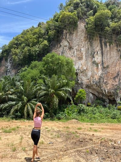 In Thung Yai, Thailand, a woman stands facing away from the camera, stretching her arms overhead. She is wearing a pink crop top and black biker shorts with white lettering. The background features a massive, rugged limestone cliff face covered in lush green foliage. Palm trees and other tropical vegetation are prominent in the foreground and midground. The ground is sandy and appears to be an unpaved area with scattered dry grass and some dirt mounds. Overhead, power lines are visible against a bright blue sky with scattered clouds.