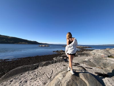 A young woman stands on rocky terrain in Nærsnes, Norway, facing away from the camera. She is wearing a black tank top, black shorts, a white shirt draped off her left shoulder, and white sneakers. The landscape features a body of water with visible islands and a forested shoreline. The sky is clear and blue. The ground is covered in rocks and seaweed.