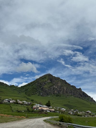 The image shows a landscape in Нижняя Мара, Russia, featuring a mountain with a rugged, rocky top and grassy slopes. A village is situated along the slope of the mountain, consisting of houses and a road. A metal guardrail is visible along the road. The sky is blue with scattered white clouds.