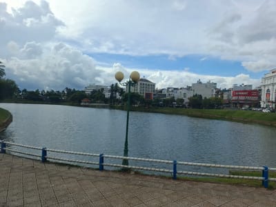 The image depicts a serene urban waterfront scene in B'Lao, Vietnam, under a cloudy sky. In the foreground, a paved walkway with a patterned surface runs alongside a calm body of water. A white and blue railing separates the walkway from the water's edge. A tall, green lamppost with two spherical yellow lights stands prominently on the walkway.
Across the water, the city skyline is visible, characterized by a mix of modern buildings and lush greenery. Notable structures include a multi-story hotel labeled "SERI HOTEL," several office buildings, and a red and white sign that reads "RIBO MART." Cars are parked and moving along a road in the background, indicating urban activity. The water reflects the overcast sky and the surrounding cityscape, creating a peaceful ambiance. The overall impression is of a well-maintained urban park area by a lake or river.
