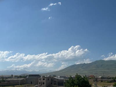 This is a wide, horizontal, eye-level shot of a town in Armenia. The sky is a bright, clear blue with scattered, fluffy white clouds. In the distance, snow-capped mountains rise, their peaks sharp and distinct against the sky. Closer to the foreground, rolling green hills form the backdrop for a collection of buildings. These buildings appear to be a mix of residential structures, some unfinished with exposed brick and rebar, and others completed with dark roofs. A lush green tree with a dense canopy stands in the right foreground, partially obscuring the view of some of the buildings. The scene suggests a sunny day in a developing or rural area.