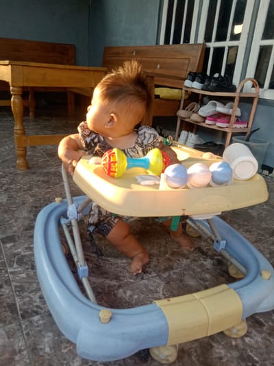 A toddler sits in a baby walker on a tiled patio in front of a building with white vertical window bars. The toddler has spiky brown hair and is wearing a patterned dress. The baby walker is light yellow and blue with a toy attached to the tray, consisting of a red and yellow ball and three blue spheres. To the left of the walker, a wooden table and bench are visible. To the right, a shoe rack holds various shoes, including black sneakers, white sneakers, sandals, and pink platform sandals. A white plastic cup or container is also present on the patio. The lighting suggests it is either early morning or late afternoon, casting warm shadows. The location is identified as Jumput, Indonesia.