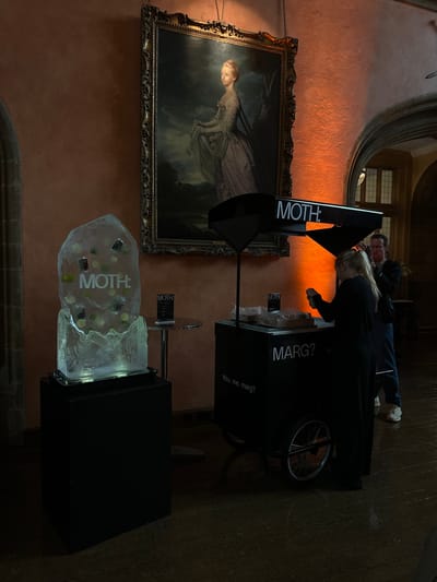 In a dimly lit, opulent interior, likely within the historic confines of Chichester, United Kingdom, a modern refreshment setup contrasts with antique surroundings. Dominating the background is a large, ornate framed oil painting depicting a noblewoman in period attire, her image illuminated by warm, orange-toned lighting. To the left of the painting, a substantial block of ice, carved into a free-standing display, contains embedded lime slices and small dark objects, possibly pre-mixed drink components. The ice sculpture is branded with the white logo "MOTH:".

In the mid-ground, a dark, mobile refreshment cart stands to the right of the ice display. It is also branded with "MOTH:" on its canopy and "MARG?" on its lower front panel, with further text below reading "You, me, marg?". Two individuals are engaged with the cart. A woman, with her back to the camera, wearing dark clothing, is actively preparing or serving from the cart. A man, wearing glasses and a darker top, stands slightly behind her, appearing to converse or assist. The cart has two visible spoked wheels.

The environment features textured, reddish-brown walls, suggesting a building with historical architecture, possibly a museum, gallery, or grand house. The floor is a dark, reflective wood, suggesting it may be wet or polished. A smaller, circular table with a black top is present between the ice sculpture and the refreshment cart, holding a menu or information card also branded "MOTH:".