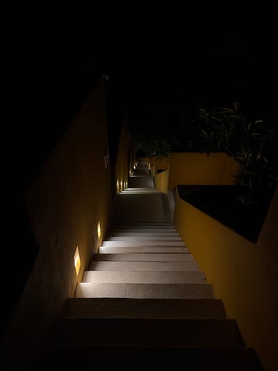This is a nighttime scene in Puerto Marqués, Mexico, featuring a partially illuminated walkway. The walkway is flanked by a series of vertical concrete structures or pillars that are lit from below by embedded, square-shaped lights. The pillars extend towards an overhanging structure, possibly a building or covered passageway, which casts a large shadow. Further in the distance, the walkway continues with additional in-ground lights illuminating the path. To the left, there's a dark area with vegetation or foliage, blending into the darkness. The foreground of the image is mostly dark. No people are visible. The overall impression is that of an outdoor architectural space with intentional lighting for ambiance and guidance.