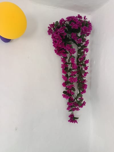 The image shows a close-up view of a corner where two white walls meet. A cascade of artificial pink flowers with green leaves hangs from the top right corner of the frame, extending downwards. To the left, a bright yellow balloon is partially visible, with a purple object beneath it. The wall appears textured and slightly uneven. There is no indication of the city of Койлык, Kazakhstan in the image.