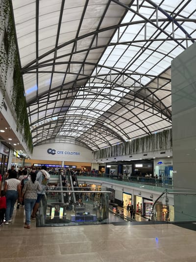 This image depicts the interior of Centro Comercial Cacique, a multi-level shopping mall located in Bucaramanga, Colombia. The primary architectural feature is a vaulted, translucent roof structure supported by a dark metal truss system, allowing natural light into the space. Suspended green plants are visible along the upper perimeters of the interior. The flooring is composed of reflective light-colored tiles.
A moderate crowd of individuals is present across multiple levels of the mall, engaged in general pedestrian movement. People are observed walking and utilizing multiple sets of escalators to transition between floors. In the mid-ground, a prominent "Cine Colombia" sign with its double-C logo indicates a cinema complex on an upper level. Below this, two sets of escalators facilitate vertical traffic. Retail storefronts line the walkways on all visible levels, including a Samsung store and a Coopetran branch on an upper floor. Digital display kiosks are positioned on the lower visible level near the escalators. Glass railings delineate the walkways and escalator openings on the upper levels.