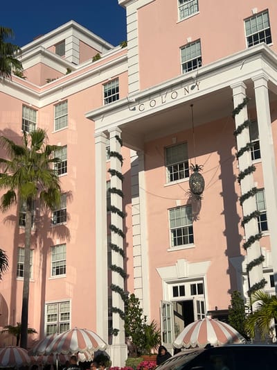 A light pink building with white columns and window frames, featuring the word "COLONY" vertically on one side. A decorative pineapple hangs under an arched entrance, casting a shadow. Palm trees are prominent in the foreground. White and pink striped awnings are visible along the building's side. Greenery adorns the building and surrounding area, set against a clear blue sky.