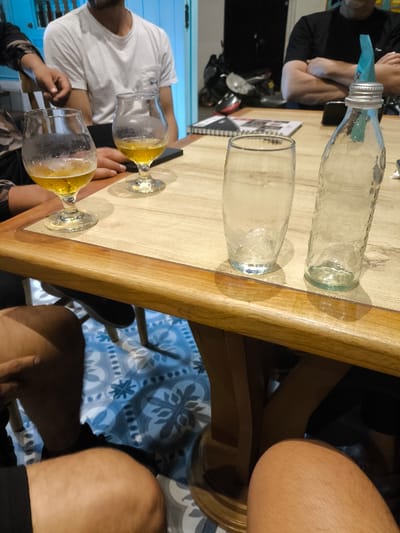 This image depicts an indoor scene within a commercial establishment in Timaná, Colombia, focusing on a wooden table occupied by multiple individuals and various beverage containers.

**People:** Four individuals are partially visible. In the immediate foreground, two sets of bare human legs are positioned under the table. Above the table, on the left, an individual wearing a white t-shirt, with a visible right arm resting on the table, is present. To their left, a person wearing a dark, patterned long-sleeved garment has a hand resting on the table. On the right, an individual in a black t-shirt sits with arms crossed on the table, their head partially cropped.

**Objects:** The central object is a rectangular wooden table with a lighter top surface and a darker wooden border. On the table are two stemmed glass beer glasses, each containing a residual amount of amber liquid and exhibiting condensation. One glass is approximately one-third full, the other less so. Additionally, there is one empty, clear, textured glass tumbler showing condensation, and one empty, clear, textured glass bottle fitted with a metallic screw cap and a light blue pourer insert. A dark-colored smartphone lies face-down on the table. In the background, a partially visible spiral-bound notebook or menu is on the table.

**Environment:** The floor beneath the table is tiled with a blue and white geometric and floral pattern. The background on the left features a light blue wall or door frame. The background on the right is dark, revealing the partial outlines of what appears to be a parked motorcycle or scooter.

**Actions/Interactions:** The individuals are seated around the table, suggesting a social gathering. The presence of multiple beverage containers indicates ongoing or recently concluded consumption of drinks.