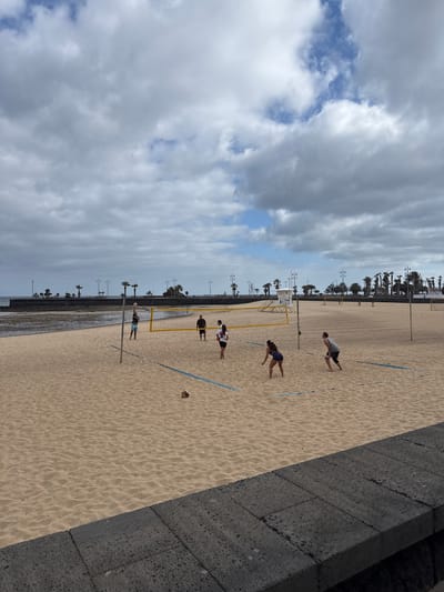 The image shows a beach volleyball game in progress on a sandy beach. The location is likely Playa del Reducto, Arrecife, Spain, given the presence of palm trees along the beach, the typical volcanic rock wall, and the distinctive appearance of the beach.

Several people are actively engaged in the game. A volleyball net divides the playing area. A volleyball is visible near the right sideline. Cloudy skies and the sea are visible in the background. Streetlights line the beach along the water.
