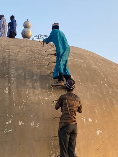 The scene depicts four male individuals on a weathered light-brown concrete dome structure in Barako, Nigeria, under a clear blue sky. Two men are actively ascending an integrated metal ladder on the dome's curved surface. The lead climber, viewed from behind, wears a teal robe, dark trousers, and light-colored sandals, gripping the ladder rungs with both hands. Directly below him, a second man, also viewed from behind, wears a brown and tan horizontally striped short-sleeved shirt, dark trousers, and a patterned cap, also gripping the ladder. Both climbing individuals wear distinct caps. At the dome's summit, partially visible, are two additional men; one in a light-colored robe and another in a blue garment. The dome's surface exhibits extensive cracking and discolored patches. Two stacked metallic spherical finials are prominently positioned at the highest point of the dome.