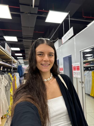 A woman with long, dark brown hair is taking a selfie in a retail store. She is smiling and wearing a white tube top and a black open cardigan. A shell necklace is visible around her neck. Behind her, racks of clothing are displayed on shelves and hanging racks. The store has a modern industrial ceiling with exposed pipes and ventilation systems, illuminated by rectangular fluorescent light panels. A sign with Vietnamese text, "PHÒNG THỬ ĐỒ," which translates to "Fitting Room," is visible hanging from the ceiling. The environment suggests a clothing store, possibly located in Phu Thuy, Vietnam, given the signage.