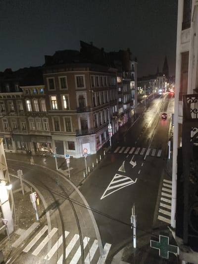 The image presents an elevated nighttime view of a street intersection in Liège, Belgium. The perspective is from a building on Rue de la Cathédrale, looking east-southeast towards the junction of Rue de la Cathédrale, Rue Hamal, and Rue des Dominicains. The prominent multi-story corner building in the left-center is identified as Rue de la Cathédrale 86.
The asphalt road surfaces are wet and reflective, indicating recent rain or damp conditions, under a dark, overcast sky. Double tram tracks curve through the foreground and midground from the lower-left towards the center-right. Multiple white zebra-stripe pedestrian crossings are visible across the intersection. Road markings include a large white triangular yield symbol and a white left-turn arrow in the mid-right portion of the road. Various streetlights illuminate the scene, casting reflections on the wet pavement.
Multi-story historical European buildings, constructed with brick or stone facades, line both sides of the streets, with numerous windows illuminated from within. A distant, tall church spire is discernible on the horizon in the background, towards the right-center. Traffic signals, street signs (including a blue square informational sign, a red circular prohibition sign, and parking signs), and white vertical bollards are present along the roadways.
One red passenger vehicle, with its rear lights illuminated, is observed in the middle distance, traveling away from the camera. Faint headlights from other vehicles are visible further down the street in the distance. No people are visible within the frame. A green cross pharmacy sign is partially visible in the bottom right corner, affixed to a building structure.