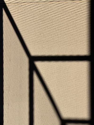 The image shows a beige textured wall with the shadows of a window frame cast upon it. The wall appears to be exterior cladding. The shadows consist of vertical and angled lines, suggesting a window with a geometric frame. There are no people or specific objects visible other than the wall and shadows. The environment is limited to the wall and shadows. The location in the city of Brasília, Brazil, cannot be determined from the image alone.