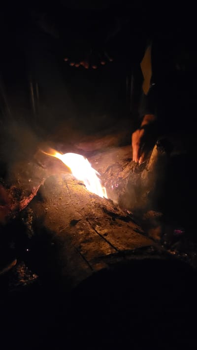 This image depicts a dark, intimate scene illuminated predominantly by a bright, crackling fire. The setting is Khir Ganga, India, suggesting a mountainous or remote natural environment, possibly a campsite or a rustic shelter.

The central subject is a vibrant fire, emanating intense orange and yellow light, which casts dynamic shadows and illuminates the immediate surroundings. Wisps of smoke are visible rising from the flames, indicating an active burn. The fire is the primary light source, emphasizing the deep darkness that envelops the rest of the frame.

Several human hands are visible, indicating the presence of people gathered around the fire. On the left side of the frame, a pair of hands with extended fingers is positioned close to the flames, suggesting they are being warmed, or are interacting with the fire, possibly stoking it or tending to something cooking. Further to the far left, lower down, another set of fingers or knuckles is faintly discernible, catching some of the fire's glow.

The ground and structures immediately surrounding the fire appear rugged and natural, likely composed of earth, rocks, or large pieces of wood. These elements are partially highlighted by the firelight, revealing their rough textures and adding to the natural, untamed aesthetic of the scene.

The activity taking place is a gathering around a fire, likely for warmth, light, and possibly cooking in the cold and dark environment of Khir Ganga at night. The atmosphere conveys a sense of primal comfort, companionship, and survival against the elements. The time of day is unequivocally night, with the fire being the sole significant light source. No visible text is present in the image.