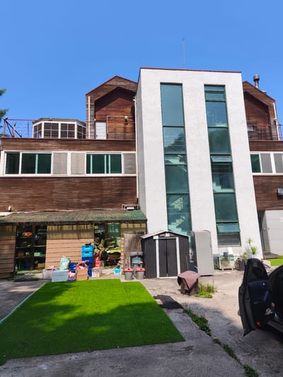 The image depicts the exterior of a building complex located in Seorak-myeon, South Korea. The complex features a multi-story structure with sections made of dark wood siding and a white stucco finish. The wooden sections have multiple windows and balconies, some with railings. The white section has tall, vertical windows that reveal an interior staircase. In front of the building, there is a paved area with a section of artificial turf laid out. Various items are scattered in the foreground and midground, including storage sheds, air conditioning units, recycling bins, boxes, and some gardening tools. A dark-colored car with its driver's side door open is parked on the right side of the image. The sky is clear and blue.