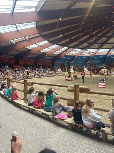 The image depicts an indoor rodeo arena in Collendoorn, Netherlands, hosting a performance for an audience. The arena is characterized by its sand-covered floor and a rustic wooden fence. A large crowd of spectators, including many children, are seated on tiered wooden benches along the perimeter of the arena, facing the performance area. Many of the children are wearing cowboy hats, suggesting a Western-themed event. In the arena, several performers are visible, including a man in a red, white, and blue shirt speaking into a microphone, and others dressed in Western attire. American flag-themed barrels are placed around the arena. The ceiling of the arena is high and features exposed wooden beams and rafters, with decorative red and yellow lights strung across it. The background of the arena is a large mural depicting a Wild West landscape with teepees, desert scenery, and figures on horseback. The overall atmosphere appears festive and engaging, with the performance drawing the attention of the seated audience.