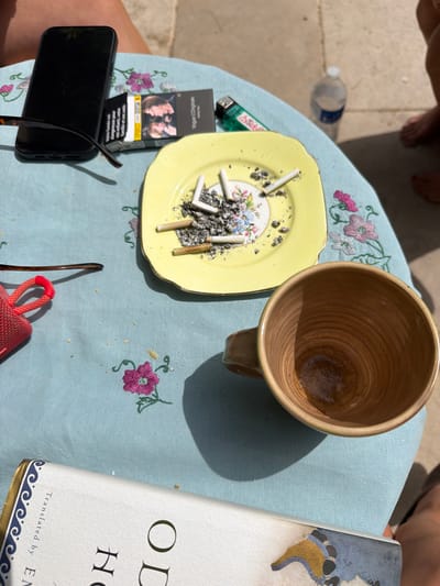 Here's a detailed description of the image:

The image depicts a casual scene, likely outdoors, on a small, light blue floral patterned table.  

**Objects:** On the table are:
*   A yellow, decorative ashtray filled with cigarette butts and ash.
*   A brown ceramic mug.
*   A pack of "Vogue L'Originale" cigarettes.
*   A green and white lighter.
*   A black smartphone with sunglasses resting on top.
*   Part of a book with "OD" and "H" visible on the cover, along with "Translated by EN".
*   A red portable speaker.
*   A plastic water bottle.

**People:** Parts of two legs are visible in the top corners of the image, implying people are seated around the table.

**Environment:** The scene appears to be outdoors, possibly a patio or garden, based on the light and shadows.

**Possible Location:** Given the implied French text on the cigarette pack, it's plausible the image was taken in France. The query mentions Saint-Victor-la-Coste, France, so it could be reasonably assumed the image was taken in this city.

**Actions/Interactions:** The scene suggests someone has been smoking cigarettes and reading, perhaps while enjoying a beverage, and listening to music.
