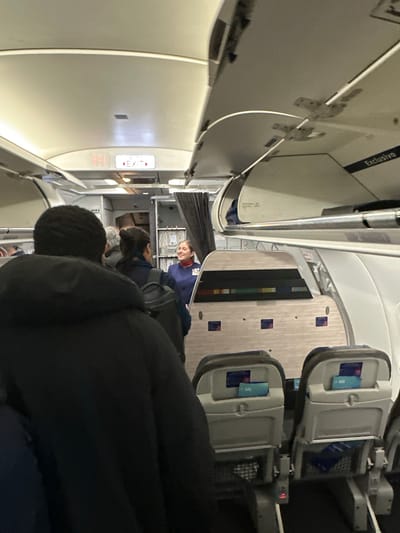 The image depicts the interior of an airplane cabin. Passengers are seated, and an air stewardess stands near the front. There is an exit sign visible, and overhead compartments line the ceiling. The scene is located in an aircraft, no evidence within the picture allows me to identify the city of Pudahuel, Chile.