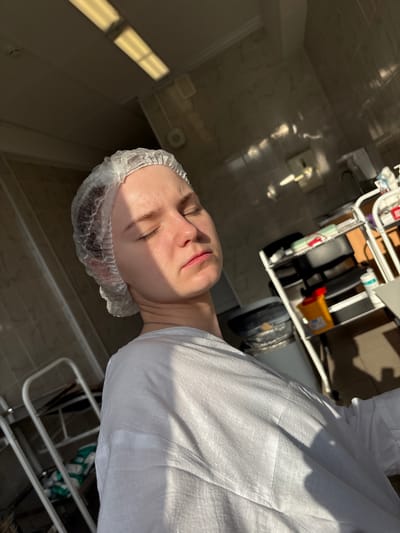 The image shows a person wearing a white disposable hair net and a white shirt. The person's eyes are closed, and they are bathed in sunlight. The background appears to be an indoor medical or laboratory setting, with tiled walls, shelving units holding medical supplies, a trash can, and fluorescent lighting on the ceiling. The overall impression is one of a moment of rest or contemplation in a clinical environment. The location is identified as Unknown, Russia.