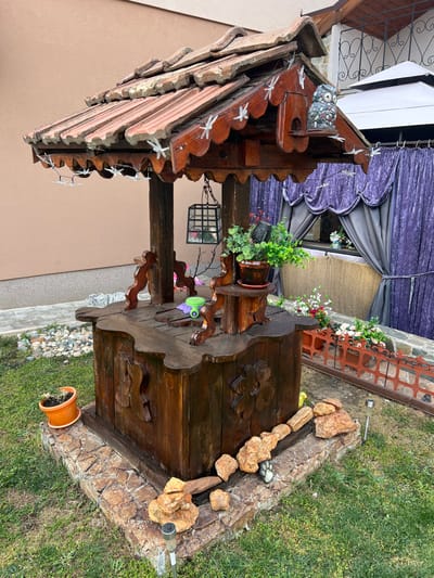 The scene depicts a decorative wooden well situated outdoors in a grassy area within the city of Mitino, Bulgaria. The well features a dark-stained wood construction with an octagonal stone-clad base. Its roof is tiled with terracotta-colored tiles, adorned with white dragonfly figures along the scalloped wooden eaves. A grey owl figurine sits on a small shelf built into the roof structure. Four carved wooden posts support the roof.
Attached to one of the posts, a small wooden shelf holds a brown pot with green foliage. A black metal lantern hangs by a chain from the underside of the roof. The flat top surface of the well displays a small green and purple object, possibly a child's toy. The well's base is surrounded by natural-colored rocks and a dark grey paw print stepping stone embedded in the grass. An orange pot with dried plant material is visible on the grass to the left.
In the background, a light brown building wall is visible to the left. To the right, rich purple velvet curtains are draped, likely part of an outdoor structure, with a white canopy or umbrella partially visible above. A low, reddish-brown decorative fence, adorned with white flowers, separates the grassy area from a paved section containing additional flowers. No individuals are present in the image.