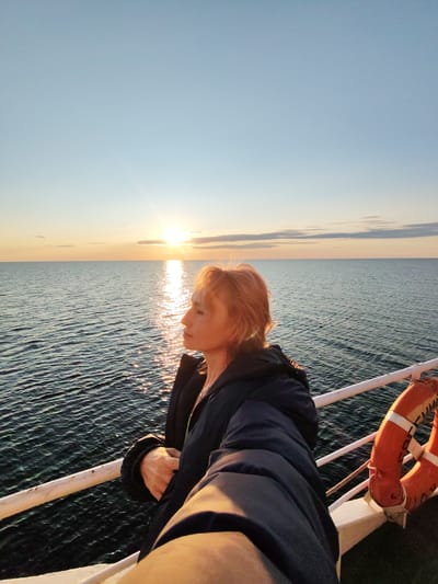 A woman with reddish-blonde hair stands on the deck of a boat, facing right and looking out at the ocean during sunset. She is wearing a dark jacket and holding her arm out, possibly for a selfie. The sun is low on the horizon, casting a warm glow on the water and the sky, with some clouds visible. A life preserver is attached to the railing of the boat on the right side of the image. The scene appears to be in the open sea.