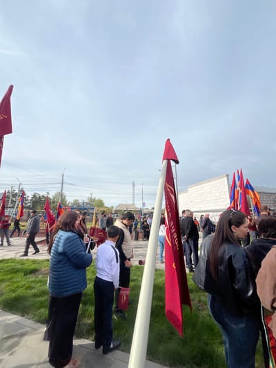 The image captures a gathering in what appears to be Akhuryan, Armenia. Several individuals stand outdoors, some holding red flags with possible Armenian inscriptions. The group includes men, women, and what seems to be students in uniform. They stand on a paved area adjacent to a grassy patch, and rows of additional flags are planted along the walkway. A building with a white facade is visible in the background. The sky is overcast, indicating daytime. The scene suggests a formal event or commemorative gathering.