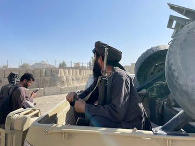 The image displays a daytime scene in Spin Boldak, Afghanistan, focusing on individuals in the rear bed of a light-colored utility or military-style pick-up truck. In the foreground, a man with a dark beard, wearing a dark turban and dark long-sleeved clothing, is seated, facing towards the right side of the frame. An M16/AR-15 variant rifle is slung over his right shoulder. His hands are visible, holding a folded green fabric object. To his left, slightly further back in the truck, another man with a beard and a patterned head covering, dressed in a light brown garment, is intently focused on a smartphone held in both hands. A dark backpack rests on his left shoulder. A tan jerry can is present in the truck bed near this individual. The background features a robust defensive barrier constructed from stacked Hesco barriers topped with razor wire, extending horizontally across the mid-ground. Beyond this fortification, a light-colored structure is visible on a distant elevated position, amidst other smaller buildings and sparse, arid vegetation, beneath a clear, bright blue sky indicative of sunny, dry conditions.